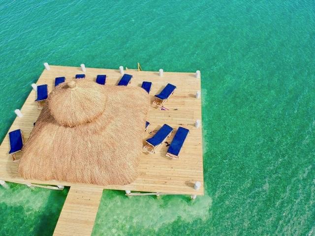 A spacious wooden dock extends over clear turquoise waters, featuring a central thatched-roof palapa. Surrounding lounge chairs with cushioned seats are positioned for relaxation. The serene environment offers an ideal setting for sunbathing and enjoying the coastal scenery.
