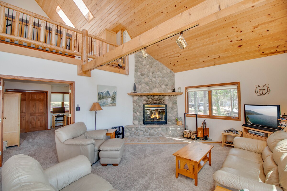 A spacious living area features a stone fireplace as a focal point, surrounded by large windows that allow natural light to fill the space. Comfortable seating options, including a light-colored sofa and armchairs, are arranged around a wooden coffee table, creating an inviting atmosphere.