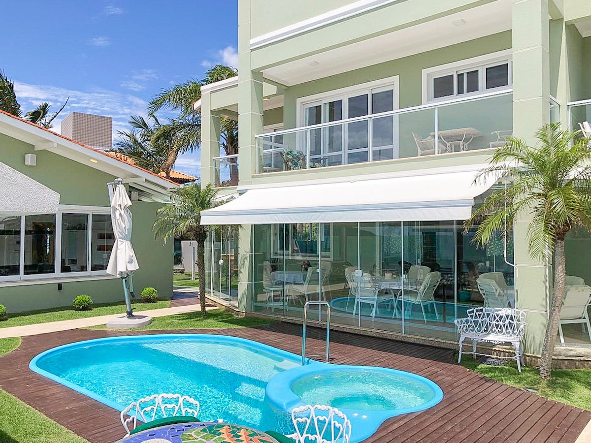 A pool area is displayed, featuring a private, turquoise pool surrounded by wooden decking. A shaded seating area with elegant white furniture is visible, complemented by palm trees and manicured shrubs. The house is painted in soft green, with large glass doors allowing natural light.