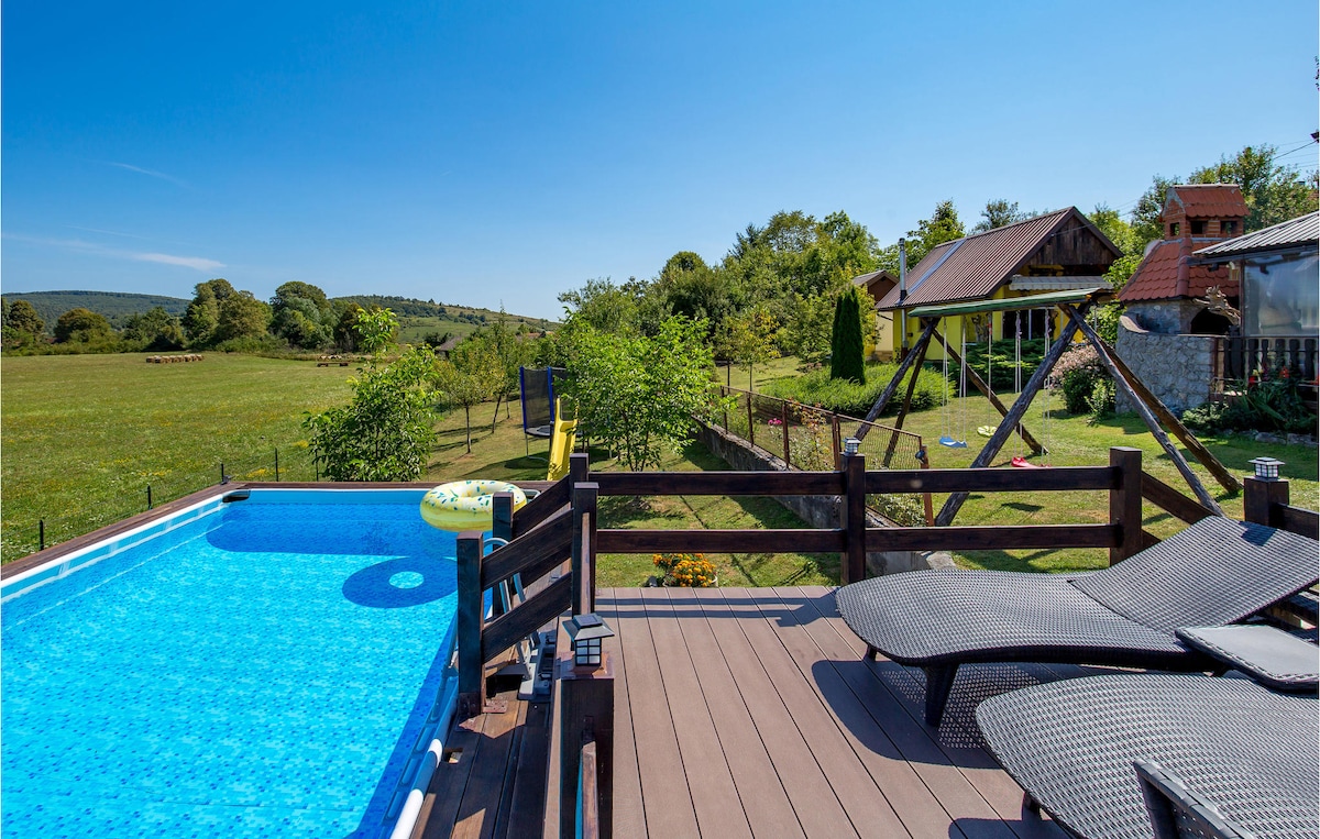 A private deck features a rectangular swimming pool surrounded by sun loungers. Lush greenery and open fields extend beyond the property, enhancing the tranquil setting. A swing set and additional structures are visible in the background, suggesting a family-friendly outdoor space.