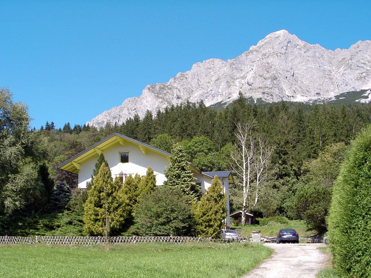A modern two-story house is surrounded by lush greenery and features a gravel driveway leading to the parking area. Towering mountains rise majestically in the background, contrasting with the vibrant blue sky.