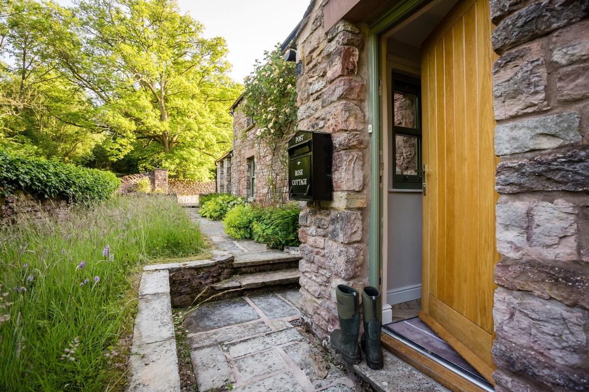 The entrance to Rose Cottage is framed by a stone pathway and lush greenery. A welcoming wooden door stands open, revealing glimpses of the interior. Two pairs of green wellington boots are placed near the doorway, suggesting readiness for outdoor adventures.