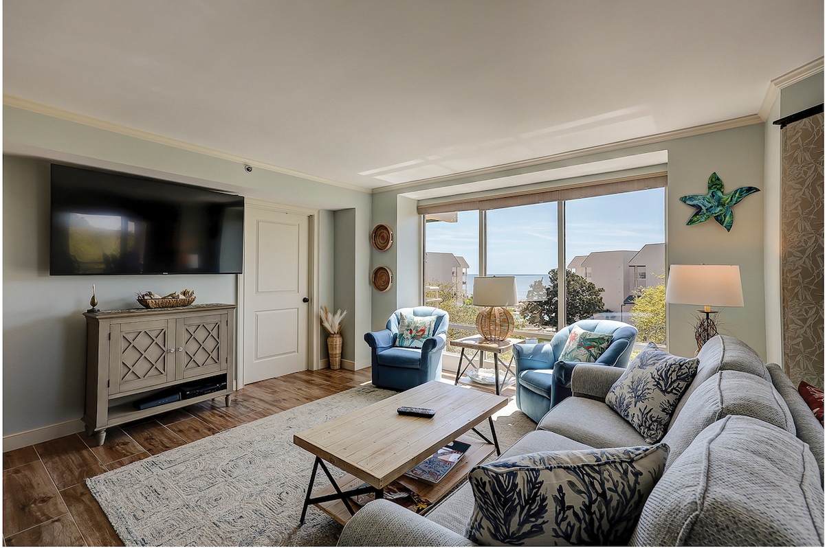 The living area is highlighted by a light grey sofa adorned with decorative pillows, accompanied by two blue armchairs. A coffee table sits in front, with a remote and a decorative bowl displayed. A large flat-screen TV is mounted on the wall, and natural light filters through a wide window offering views of the outside.