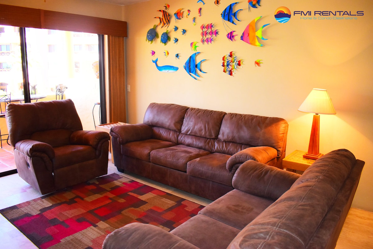 A spacious living area features two comfortable couches and a patterned area rug. A decorative wall art display of colorful fish is positioned above the seating. Natural light enters through sliding glass doors leading to a balcony, complemented by a lamp providing additional illumination.