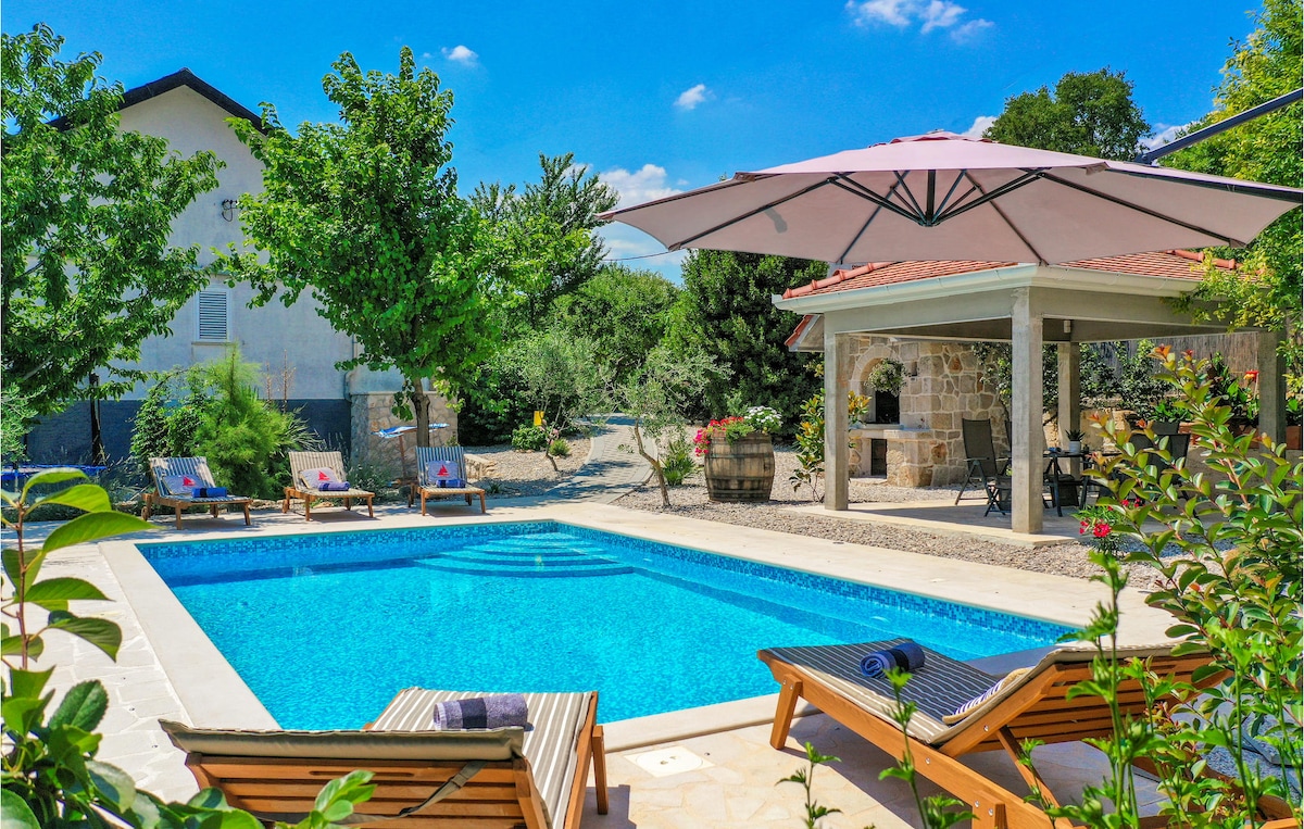 An outdoor pool is surrounded by greenery, sun loungers, and an umbrella for shade. A covered terrace is visible in the background, featuring a barbecue area. Bright blue skies and scattered clouds complement the peaceful setting.