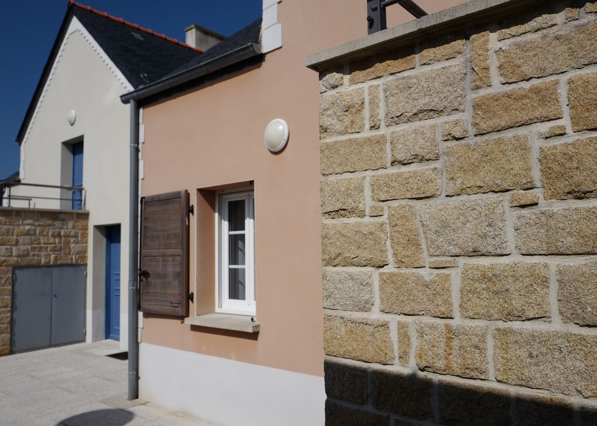 A view of a light-colored building featuring a well-maintained stone wall and wooden shutters. The simplicity of the architecture is complemented by clear blue skies, creating an inviting atmosphere outside the residence.