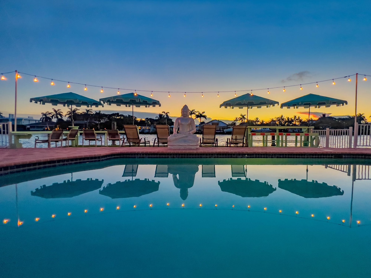 The pool area is illuminated by string lights, reflecting off the water’s surface. Umbrellas provide shade over several lounge chairs, while a statue sits peacefully at the edge of the pool, framed by a sunset sky transitioning to twilight.