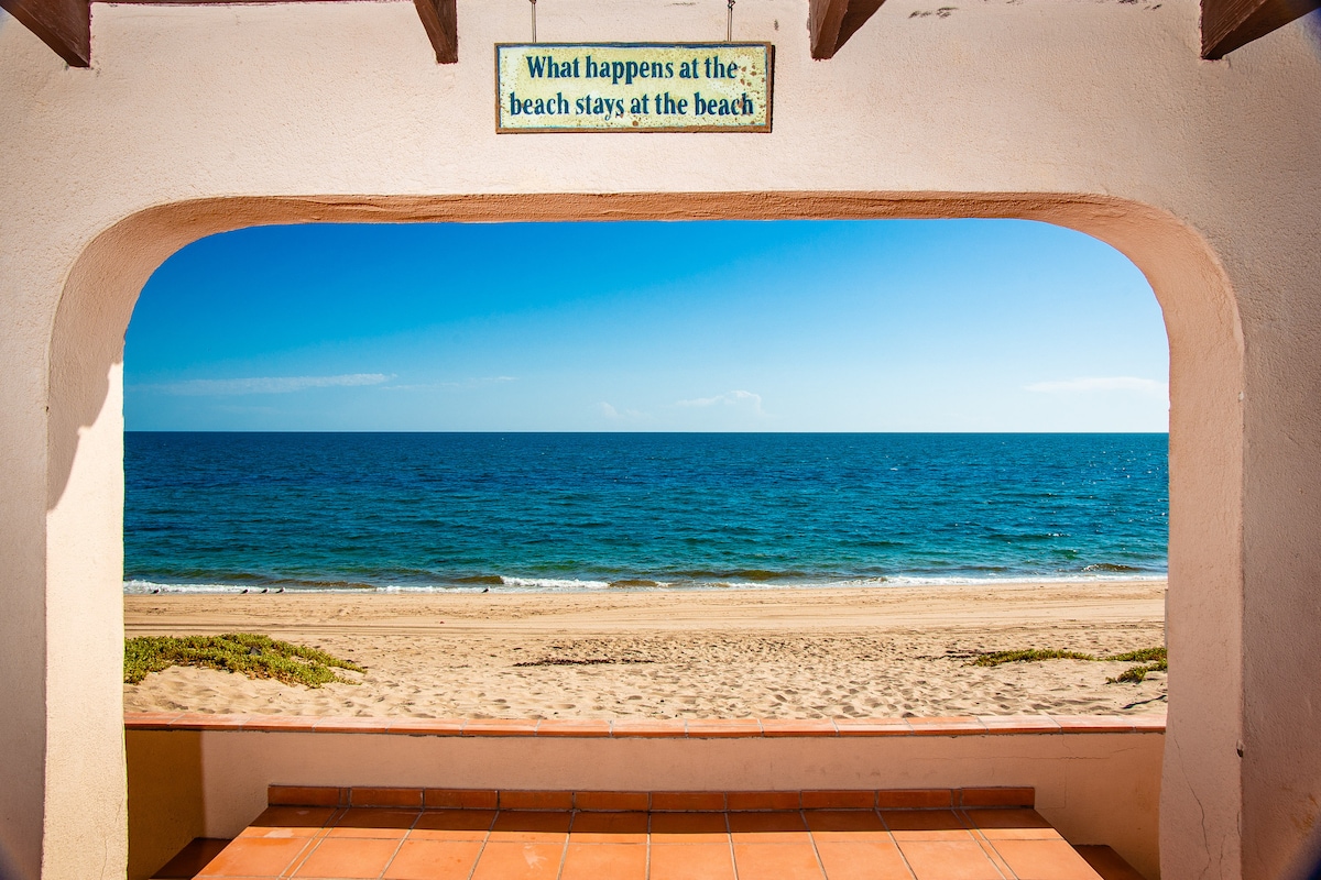 An arched opening provides a clear view of a sandy beach and serene ocean waters. The sky above is bright blue, complementing the calm sea. A tile floor can be seen beneath the arch, enhancing the beachside setting.