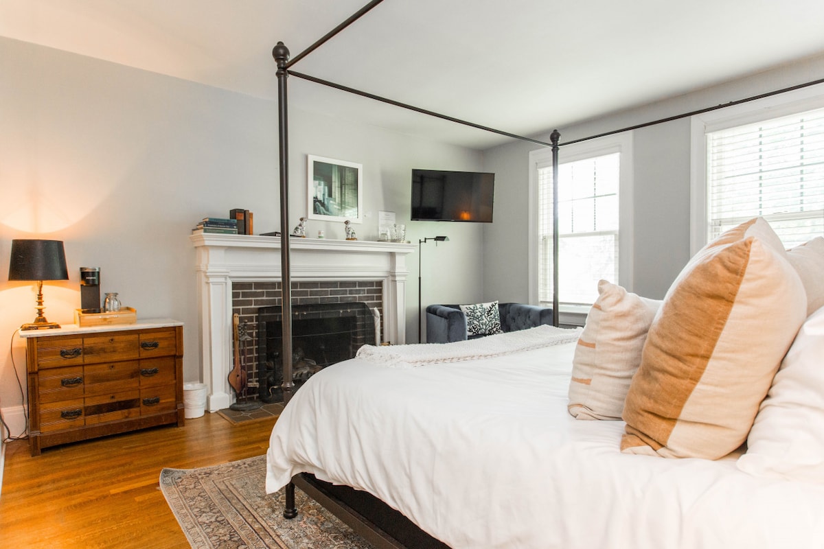 A spacious bedroom is accentuated by a king-sized four-poster bed with crisp white linens. A wooden nightstand and a dresser are positioned nearby, while a fireplace adds a touch of warmth. Natural light filters through multiple windows, illuminating the room's neutral tones.