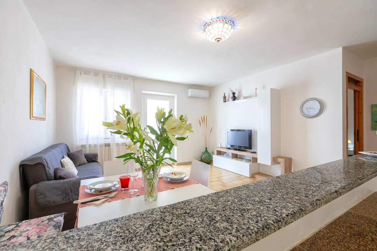 A bright living area features a comfortable sofa, a television unit, and a dining space set with plates and glasses. Large windows allow natural light to fill the room, while a decorative light fixture hangs from the ceiling, enhancing the space.