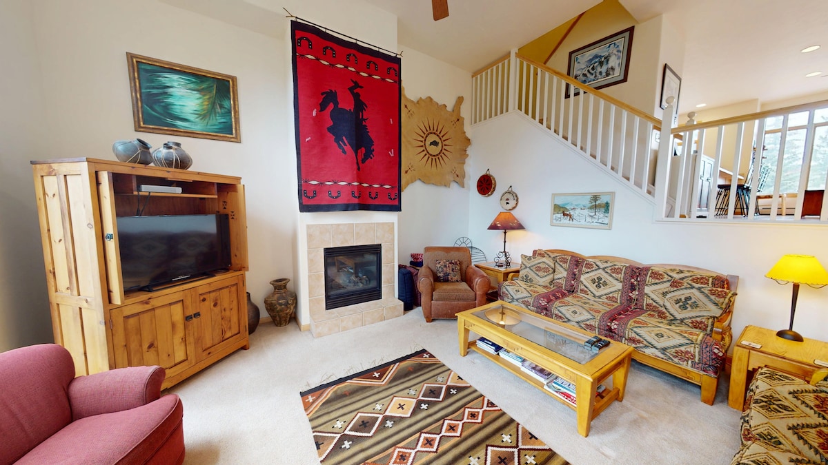A welcoming living area features a gas fireplace framed by a natural stone surround. Comfortable seating, including a deep red armchair and patterned sofas, are arranged for relaxation. Colorful artwork and a vibrant rug add character, while a wooden TV stand holds a flatscreen television.