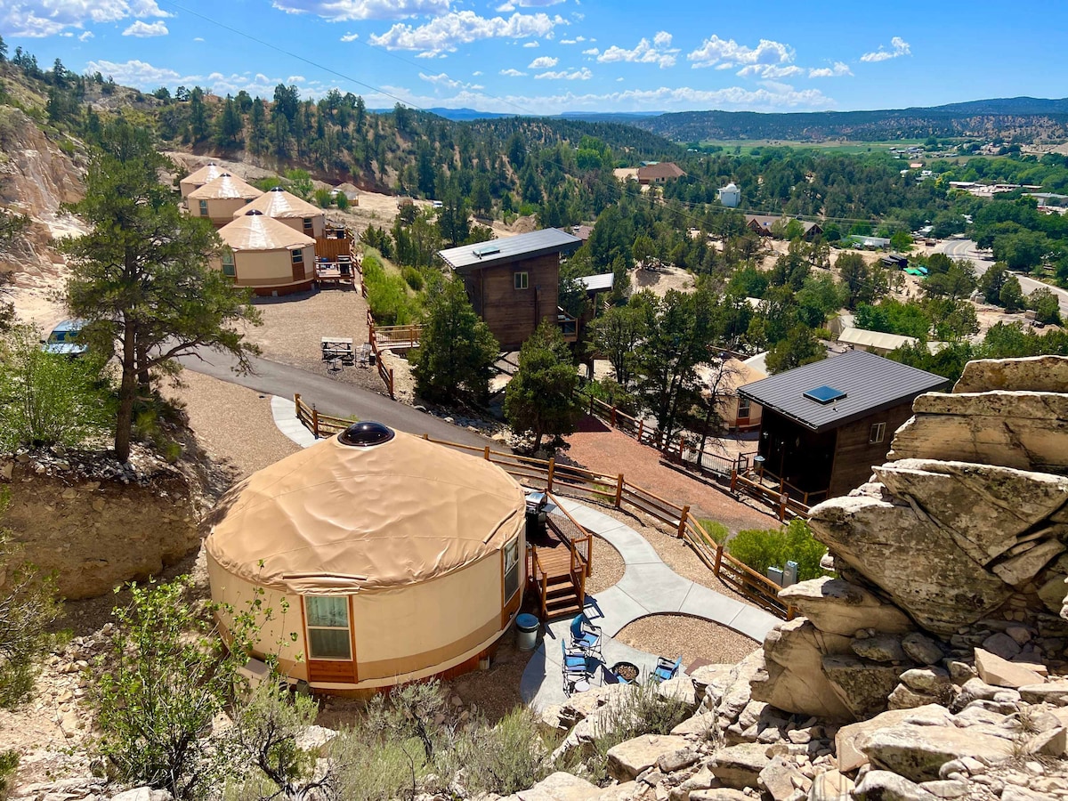 A panoramic view reveals multiple yurts nestled among trees and rocky terrain, overlooking the scenic landscape. A walking path connects the structures, while outdoor seating arrangements are positioned near the yurts, inviting relaxation. The clear blue sky enhances the tranquil atmosphere of this unique hillside setting.