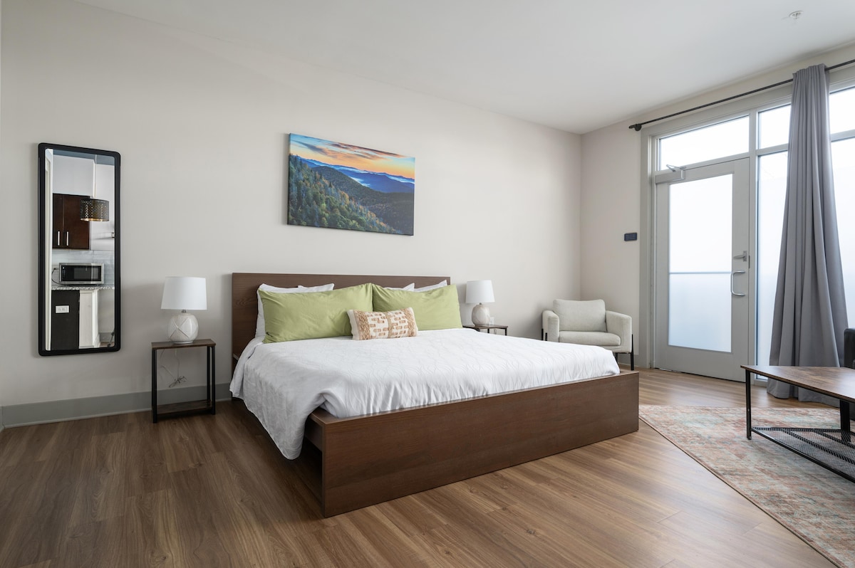 A modern studio room features a spacious bed topped with soft green pillows and a decorative throw. Natural light enters through large glass doors, complemented by a small seating area and a coffee table. A piece of landscape artwork is displayed above the bed.