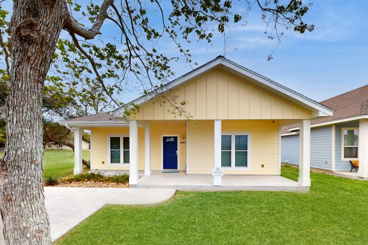 A modern beach house with a yellow exterior and blue front door is framed by a covered porch. Green grass and a large tree enhance the inviting entrance. The home is situated in a peaceful neighborhood, surrounded by open space.
