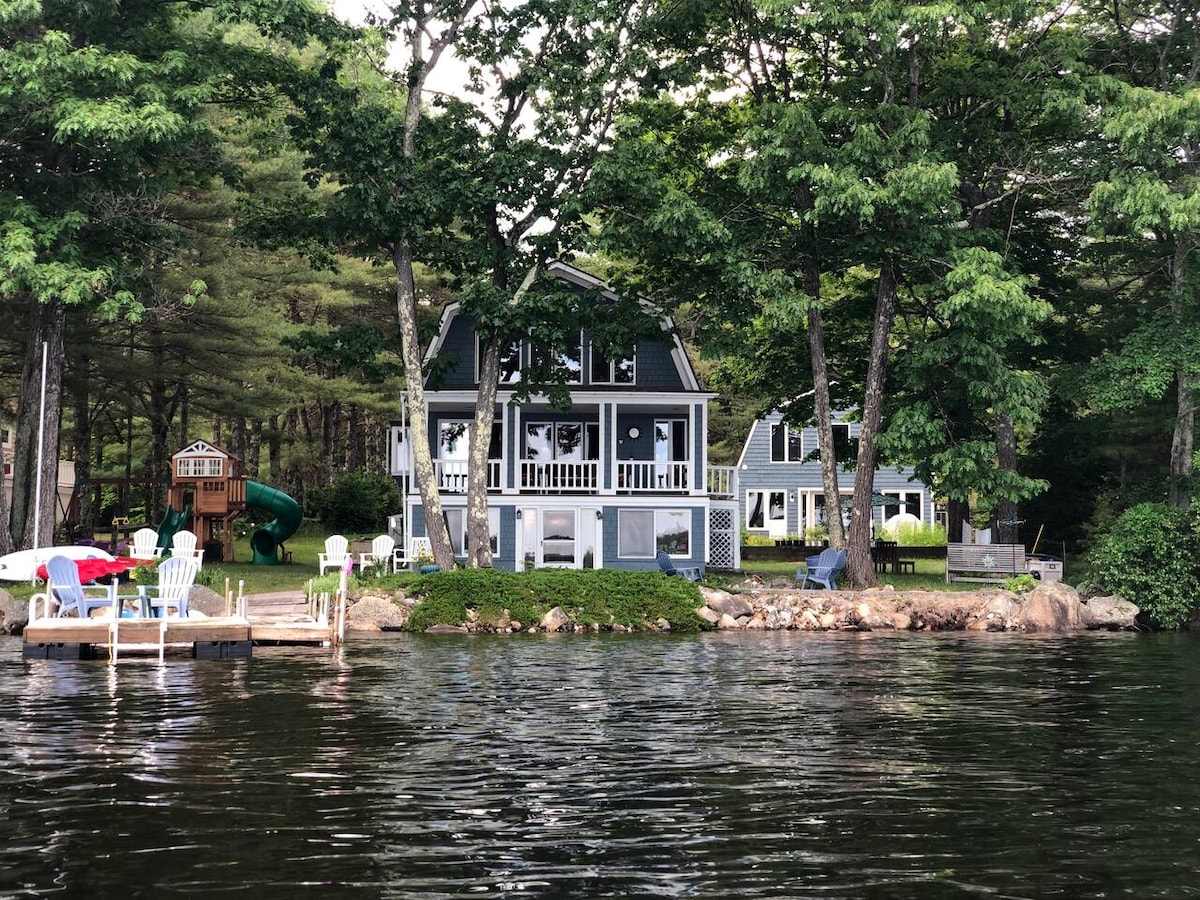 A view of two homes situated closely near the water, framed by trees. The main house features a deck with a balcony above. A children's playset is visible to the left. The calm lake reflects the greenery surrounding the property.
