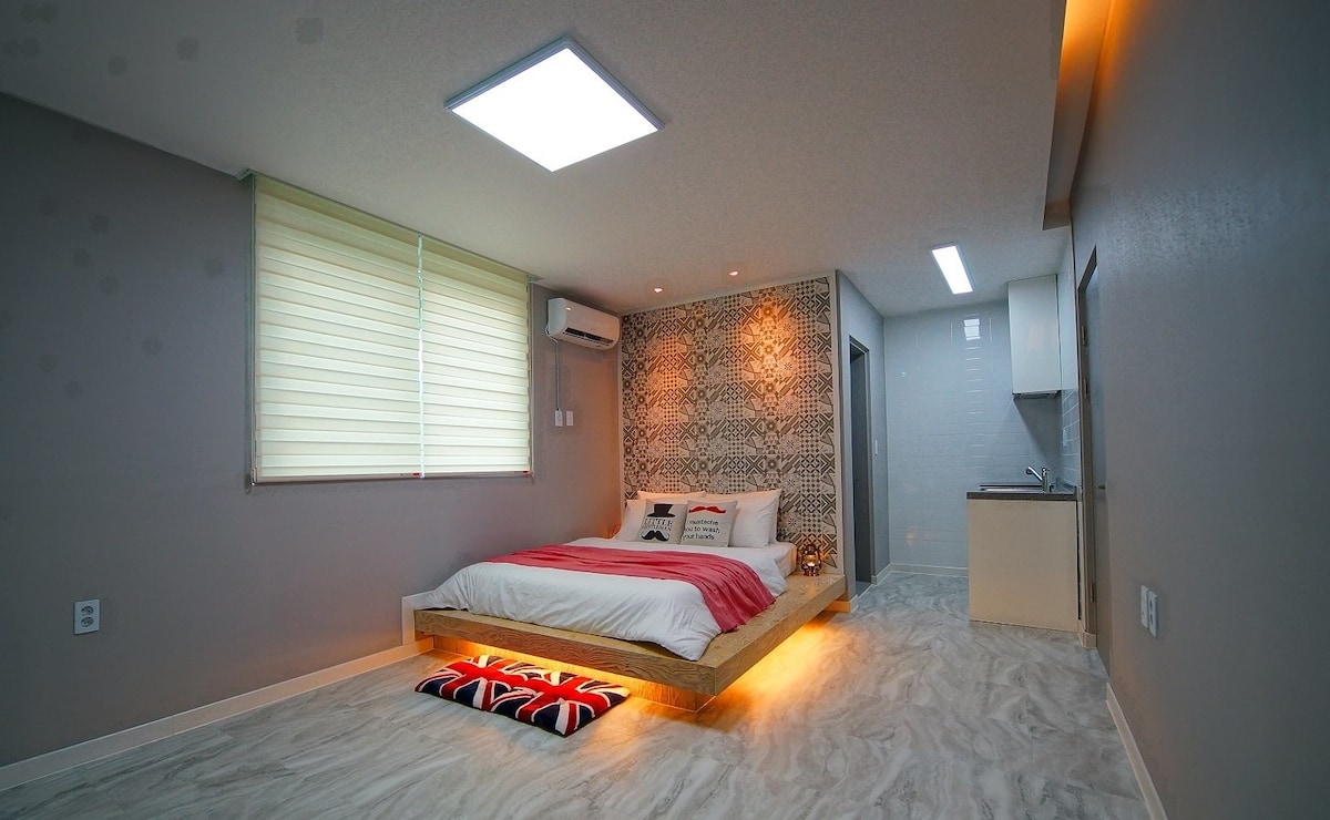 A modern bedroom features a spacious bed with a low wooden frame and decorative pillows. Natural light enters through a window covered with light shades. A kitchenette is visible in the background, along with simple decor that adds a contemporary feel.