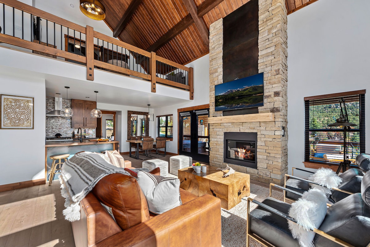 The open great room features a vaulted wooden ceiling and a stone fireplace that extends from floor to ceiling. Comfortable seating includes leather sofas and armchairs. Natural light floods the space through large windows, offering mountain views and creating a warm, inviting environment.