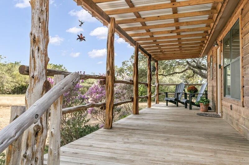 A wooden porch with rustic railings is featured, offering views of the surrounding landscape. Two comfortable adirondack chairs are positioned next to a potted plant. Natural light enhances the inviting space, while a decorative mobile adds a touch of charm.