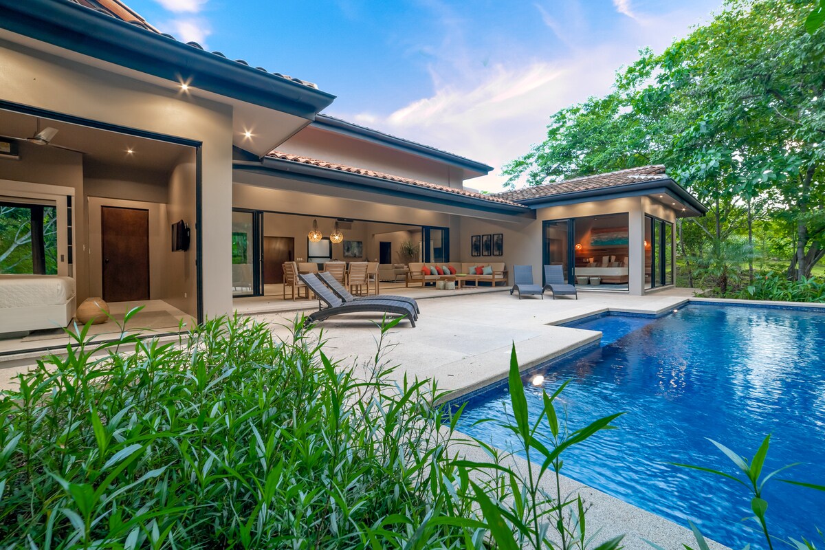 An outdoor area features a private pool surrounded by lush greenery, with comfortable lounge chairs positioned nearby. The open layout of the home is showcased through large glass doors, inviting natural light into the living space, with a view of the jungle landscape.