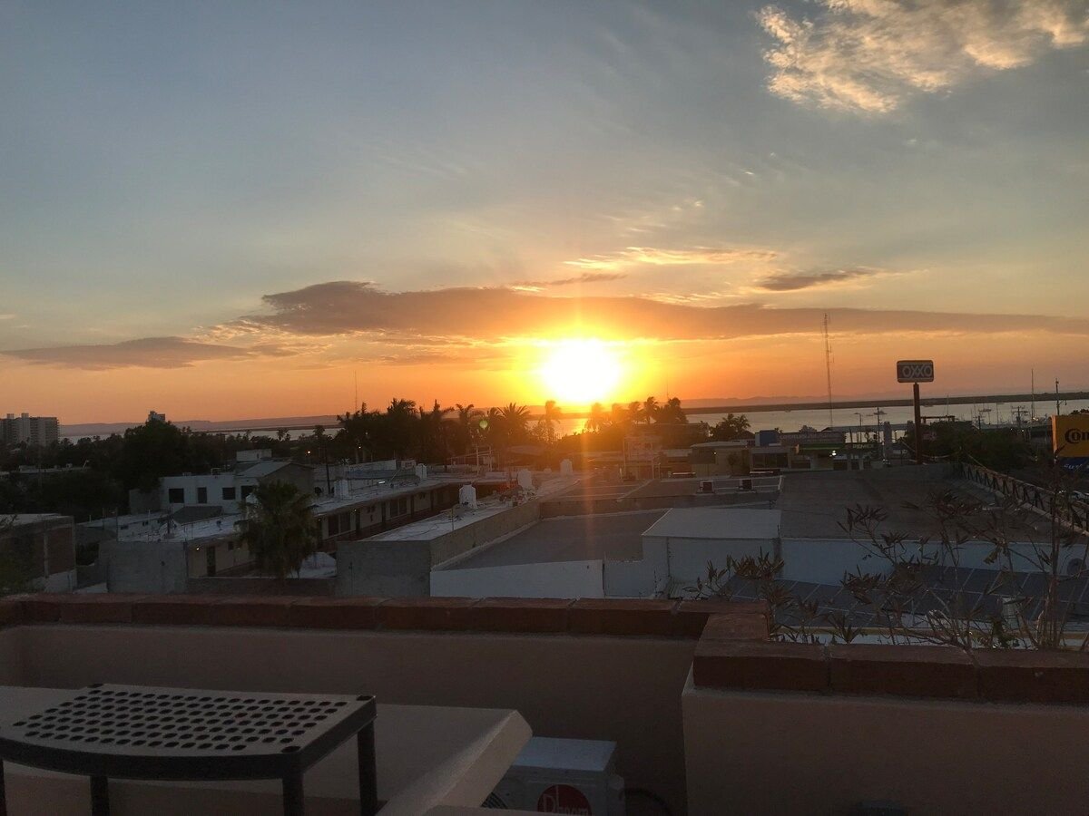 A sunset view is captured from a rooftop terrace. The sun descends toward the horizon, casting warm hues across the sky. Buildings and palm trees are visible in the foreground, with the bay outlined in the distance, offering a scenic backdrop.