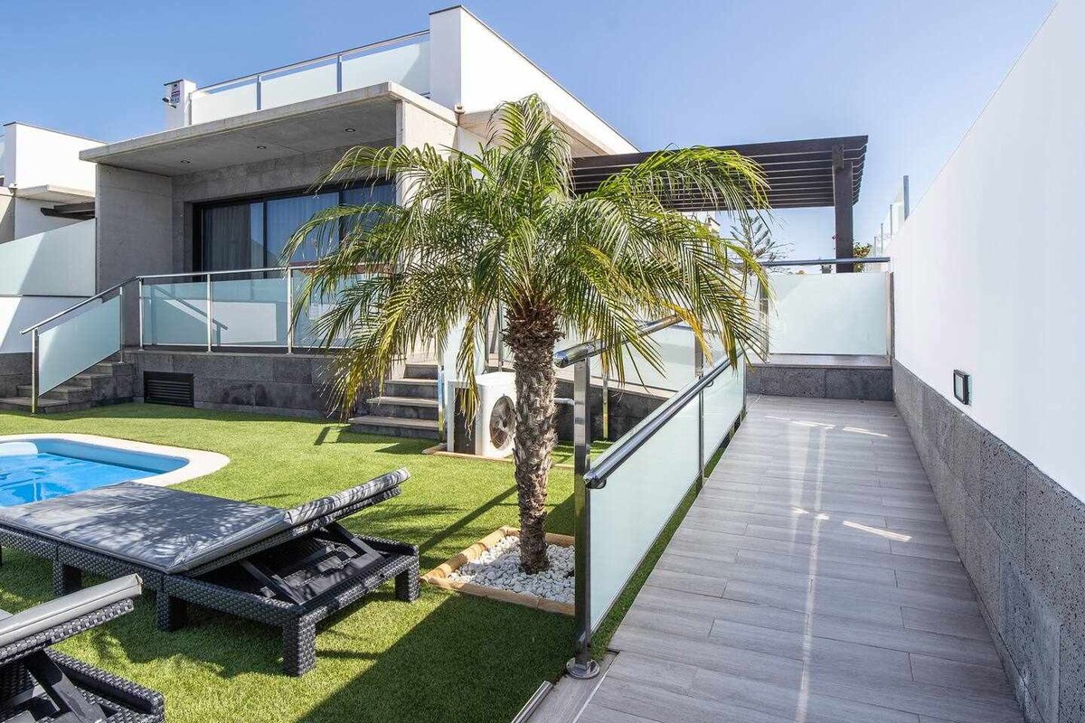 An elegant outdoor space features a private pool surrounded by manicured grass and lounge chairs. A palm tree provides a touch of greenery, while a modern path leads to a covered terrace, enhancing the open and airy design of the villa.
