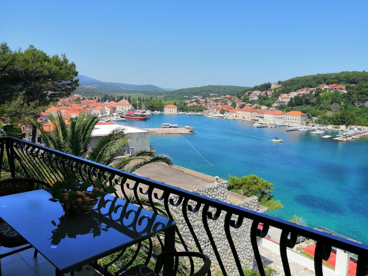 A balcony offers a panoramic view of the coastal town, featuring colorful rooftops and a serene marina. The turquoise water reflects the clear sky, while greenery frames the scene. A dining table with a fruit bowl is positioned on the balcony, inviting relaxation.