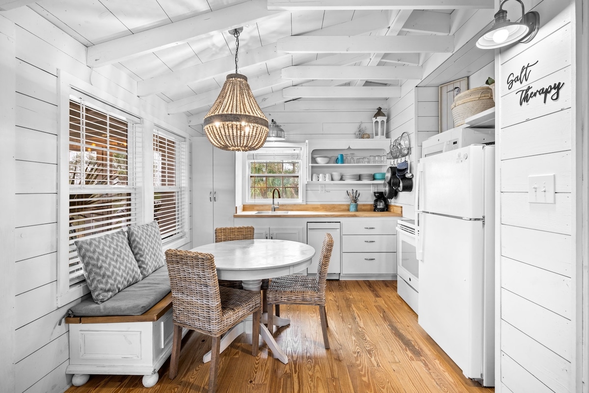 A bright kitchen area features white shiplap walls and a vaulted ceiling. A round table with four woven chairs is positioned near a bench seat. Shelves above display dishes and kitchen essentials, while a vintage light fixture hangs above the table, illuminating the welcoming space.