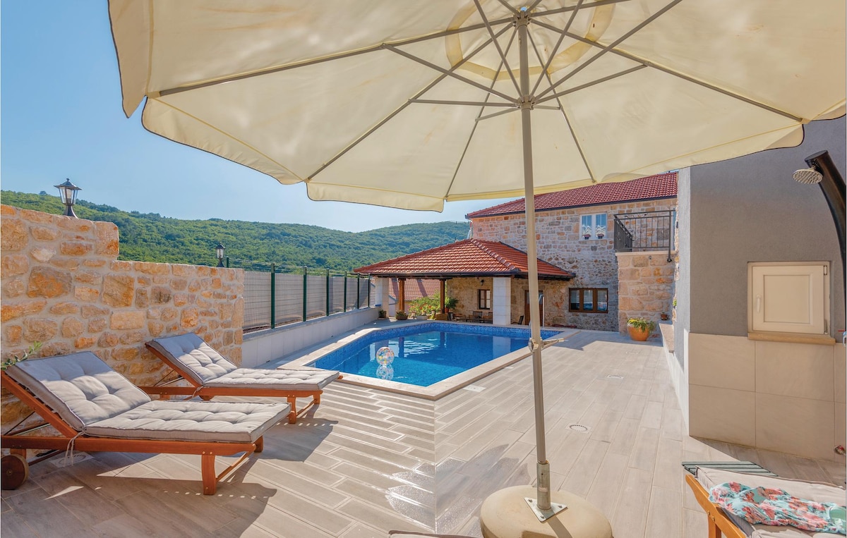 A spacious outdoor area features a private pool surrounded by a sun deck. Two stylish lounge chairs are positioned under a large umbrella, providing shade. Nearby, the stone house's rustic façade complements the scenic hillside backdrop.
