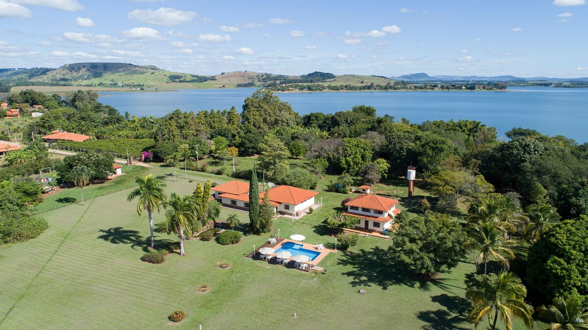 An aerial view of a spacious landscape featuring a house with a red-tiled roof surrounded by lush greenery. A bright blue pool is situated on a sunlit patio, with the expansive Represa de Chavantes visible in the background and gentle hills framing the scene.