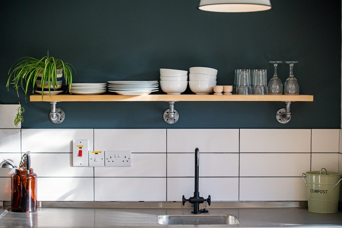 A wooden shelf is mounted on a dark wall, displaying an arrangement of white plates, bowls, and glasses. Below, a modern black faucet is positioned above a stainless steel sink. A small potted plant hangs alongside, adding a touch of greenery to the space.