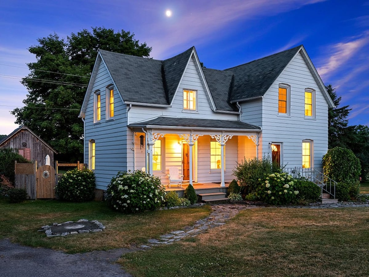 A charming vintage farmhouse stands illuminated in the evening, showcasing warm yellow lights from within. The well-kept yard features greenery and flowering plants, while stone pathways lead to the welcoming front porch adorned with decorative trim.