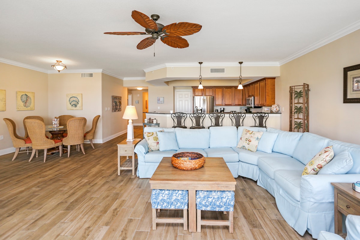 A spacious living area features a light blue sectional sofa and a wooden coffee table at its center. Rattan dining chairs and a dining table are visible in the background, with a well-equipped kitchen beyond. A ceiling fan adds a touch of comfort to the space.