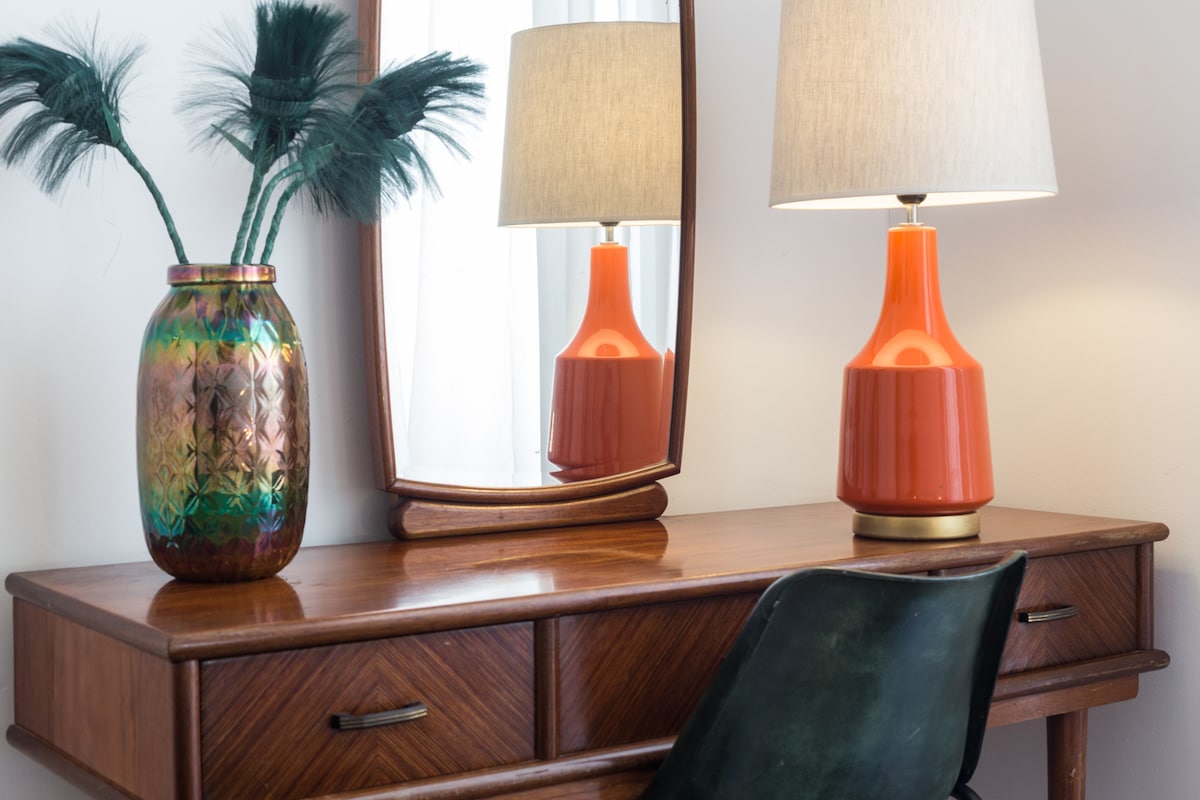 A vintage wooden desk is pictured, complemented by a round mirror above it. An orange lamp casts a warm light next to a colorful vase filled with decorative greenery. A dark green chair is positioned in front of the desk, inviting a cozy and functional workspace.