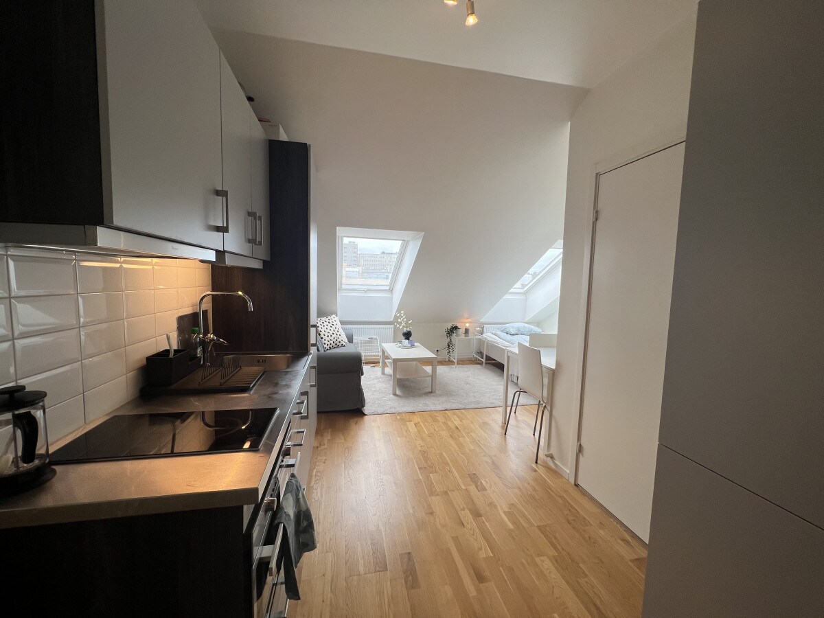 A modern kitchen space is visible with dark cabinetry and a sleek countertop. A cozy seating area includes a light-colored sofa and a coffee table, illuminated by natural light through slanted windows. A dining table for two is positioned nearby, complementing the studio layout.