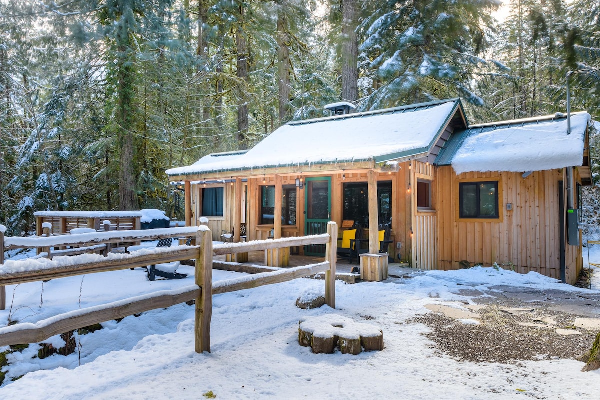 A charming cabin is nestled among trees, surrounded by a snowy landscape. The wooden exterior features large windows, allowing for natural light, while a deck with seating overlooks the serene setting. Snow covers the ground, providing a tranquil winter ambiance.