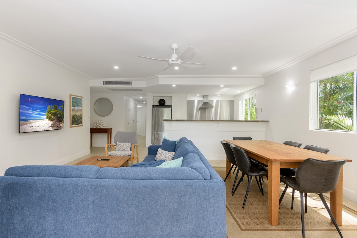 A spacious open-plan living area is featured, showcasing a comfortable blue sofa facing a flat-screen TV. A wooden dining table with black chairs is positioned nearby, with natural light coming through the nearby windows. The kitchen area is visible in the background.
