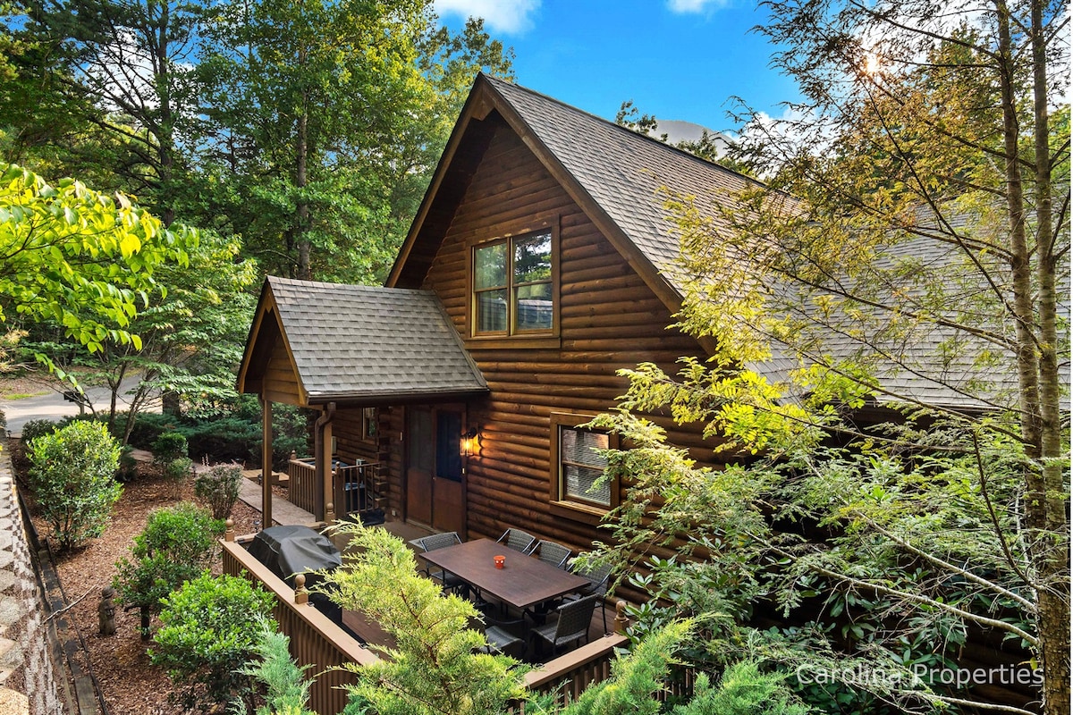 A charming log home is surrounded by lush greenery and trees, showcasing a welcoming entryway. A spacious outdoor patio features a table set for dining, inviting relaxation in a natural setting.