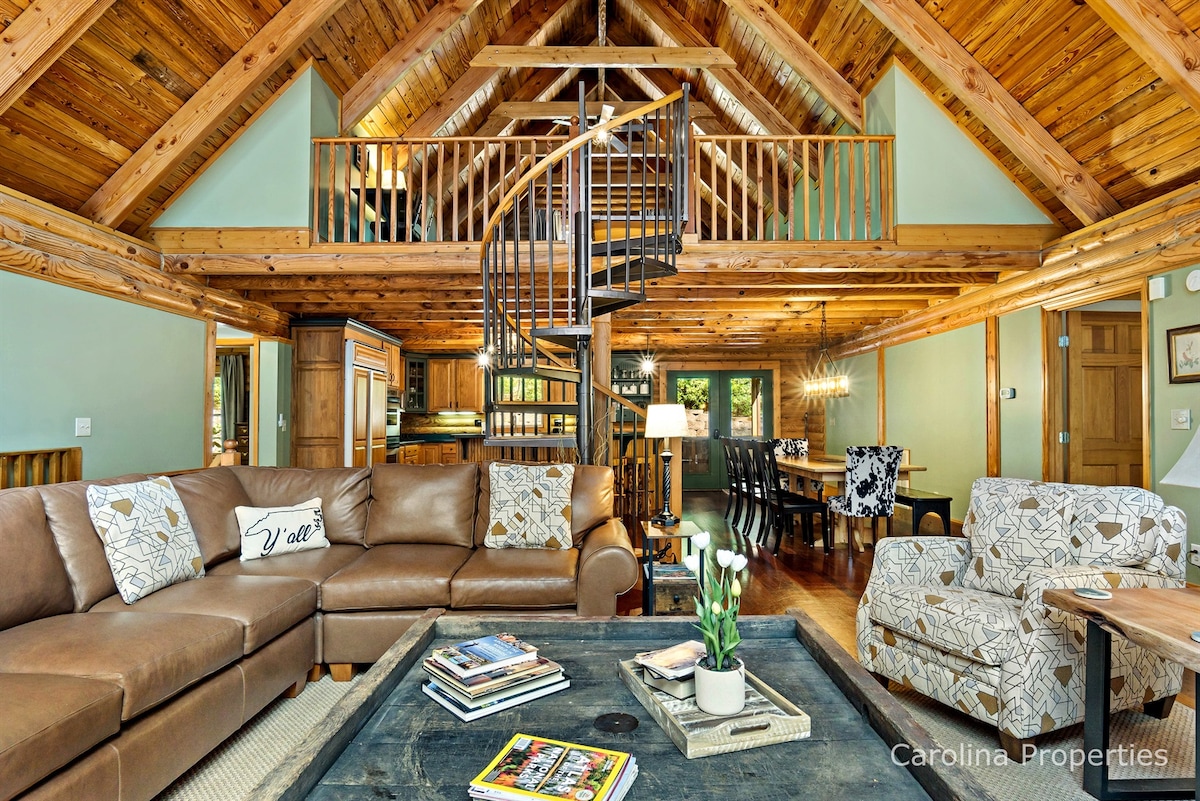 The main living area features a spacious sectional sofa with decorative cushions, arranged around a coffee table. A spiral staircase leads to an open loft. The timbered ceiling showcases a warm colour palette, while a dining area is visible in the background.