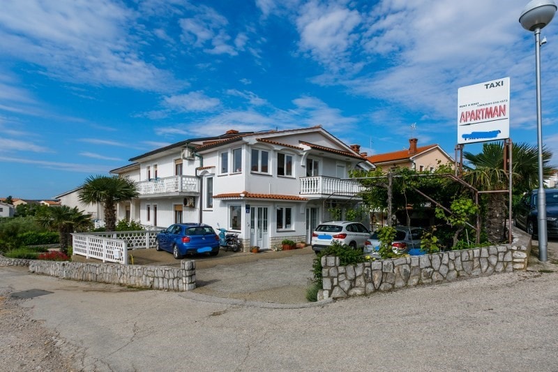 The exterior of the apartment building is characterized by a two-story white façade with a terracotta roof. A garden area with greenery adds appeal, while parking spaces are visible in front. A sign indicating taxi services can be seen nearby, contributing to convenient access.