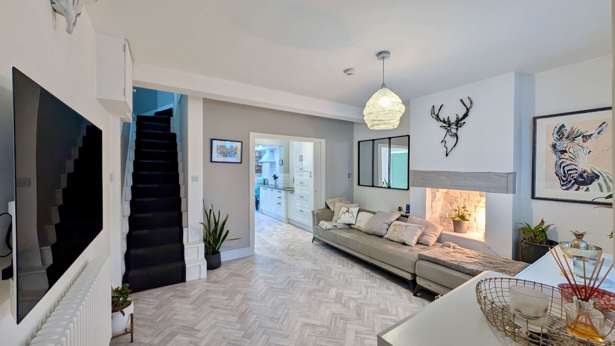A spacious living room features a modern, oversized sofa, complemented by light cushions and warm throws. An elegant fireplace is visible alongside a stylish wall-mounted mirror and a framed artwork of a zebra. A staircase leads to the upper level, enhancing the room's open feel.