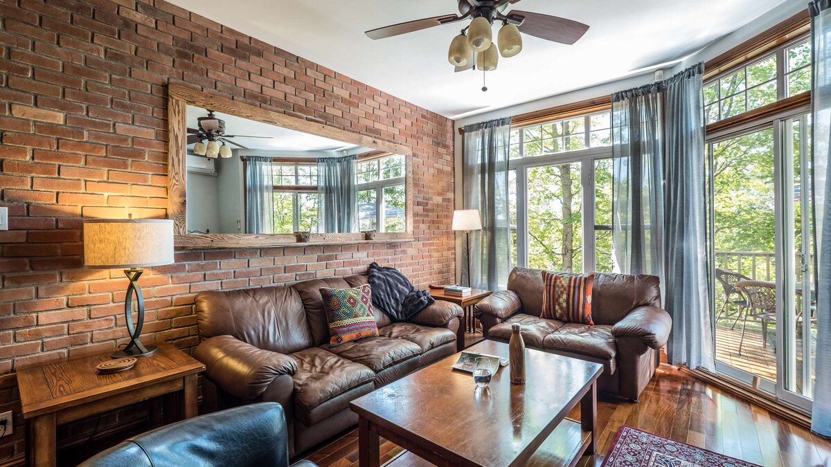 A cozy living area features a brown leather sofa and armchair set, complemented by a wooden coffee table. Natural light enters through large windows adorned with sheer curtains, illuminating the exposed brick wall and the mirror above. A ceiling fan hangs, adding comfort to the space.