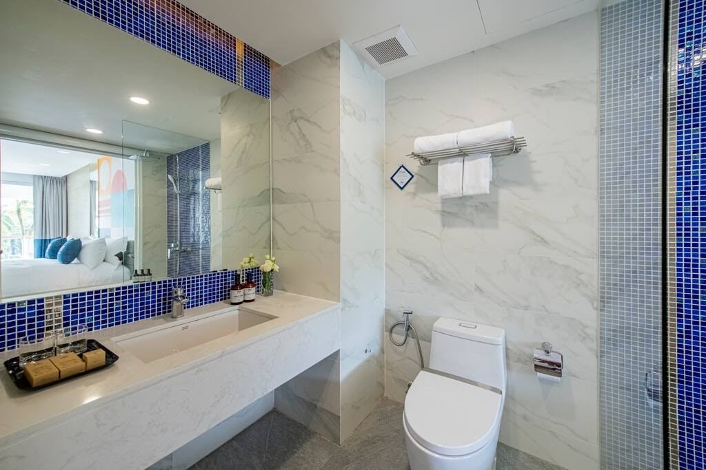 The bathroom is designed with a modern aesthetic, featuring marble walls and a spacious vanity. A shower area with glass doors is visible, alongside a toilet and neatly arranged towels on a shelf. A decorative arrangement of toiletries is placed on the vanity.