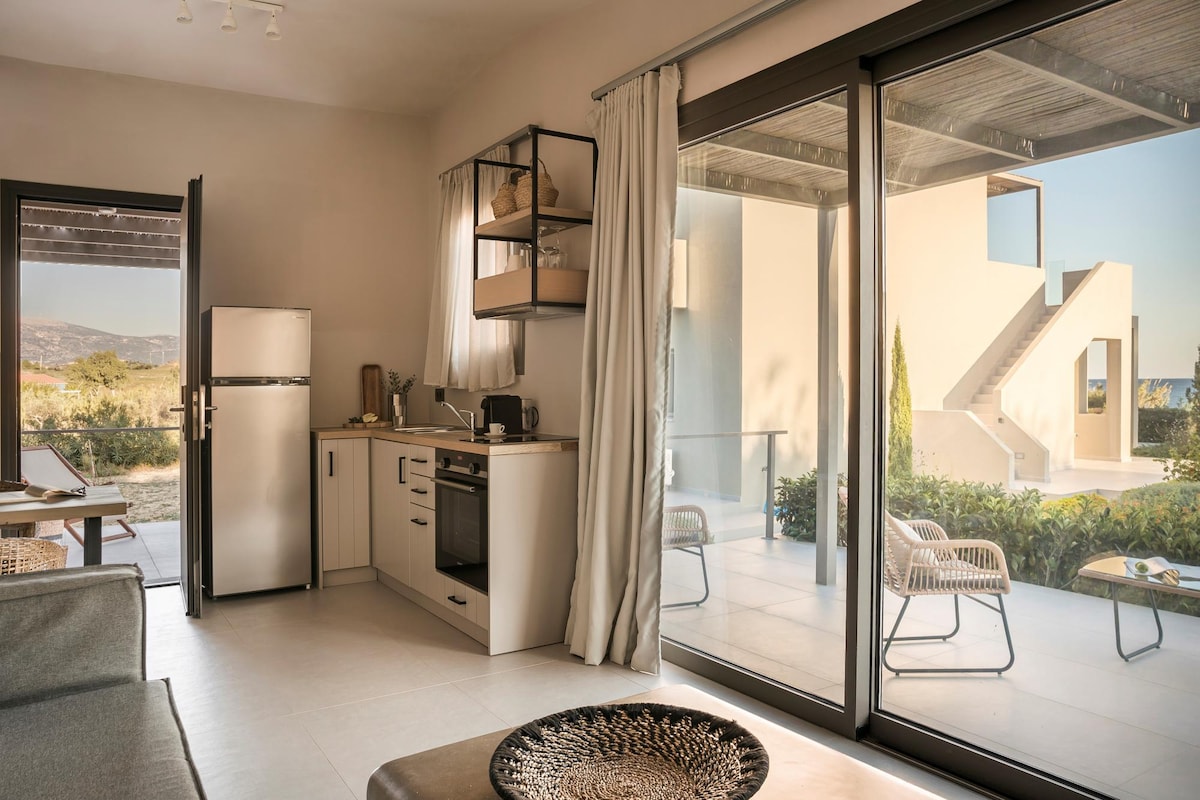 An open-concept kitchen and living area are visible, featuring a sleek refrigerator and stove. Large glass doors allow natural light to fill the space, offering views of the outdoor patio and garden. Neutral tones create a calming atmosphere in this well-designed suite.