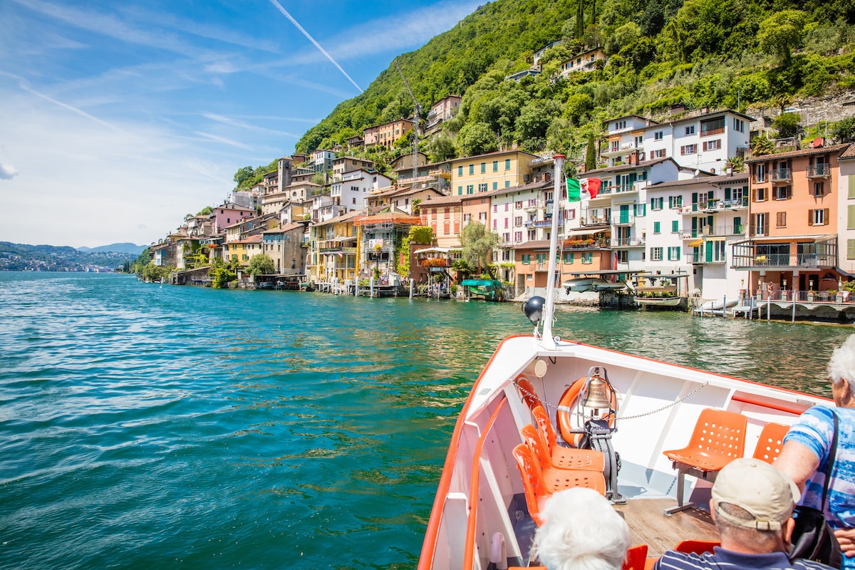 A scenic view of colorful buildings lining the lakeshore, seen from a boat. The clear blue water reflects the vibrant facades, while a lush green hillside serves as a backdrop. Outdoor seating is visible on the boat, inviting a relaxing journey along the lake.