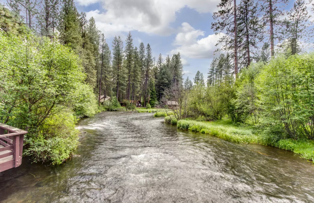 A scenic view of the Metolius River is presented, surrounded by lush greenery and tall evergreen trees. The water flows gently, reflecting the sky and vegetation. A wooden bridge can be seen in the foreground, connecting nature and facilitating access to the tranquil setting.