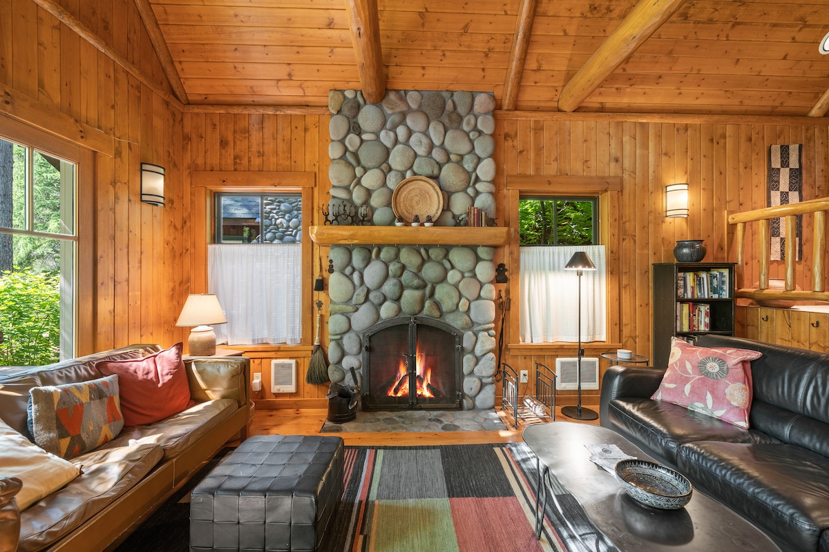 A welcoming living area features a stone fireplace at its center, surrounded by wooden walls and beams. Two windows allow natural light to illuminate the room, while a variety of seating options and decorative elements create a comfortable and inviting space.