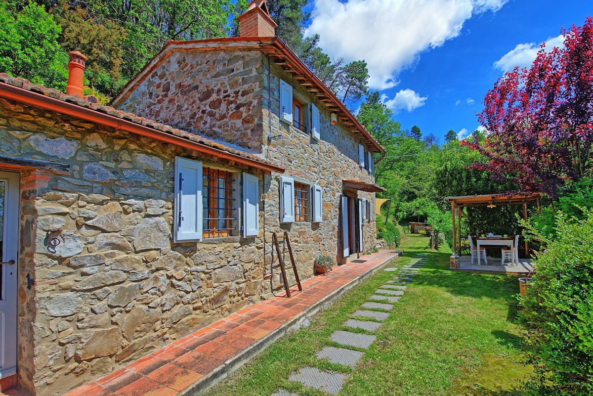 The exterior of the stone villa showcases a charming design with a sloped roof and multiple windows. A well-maintained garden features a pathway leading to a covered outdoor dining area under a wooden pergola. Lush greenery and trees surround the property.
