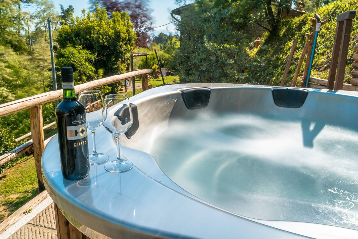 A Finnish tub, designed for five people, is shown with bubbling water and LED illumination. Two glasses are positioned beside a bottle of wine on the edge. The surrounding garden is visible, featuring lush greenery and a serene atmosphere.