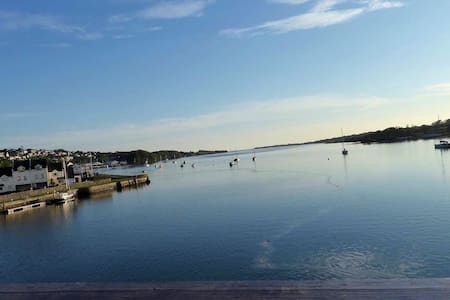 The image presents a serene view of the Menai Straits, showcasing calm waters reflecting clear blue skies. Boats can be seen gently bobbing in the distance, while the shoreline features quaint buildings and greenery on the opposite bank.