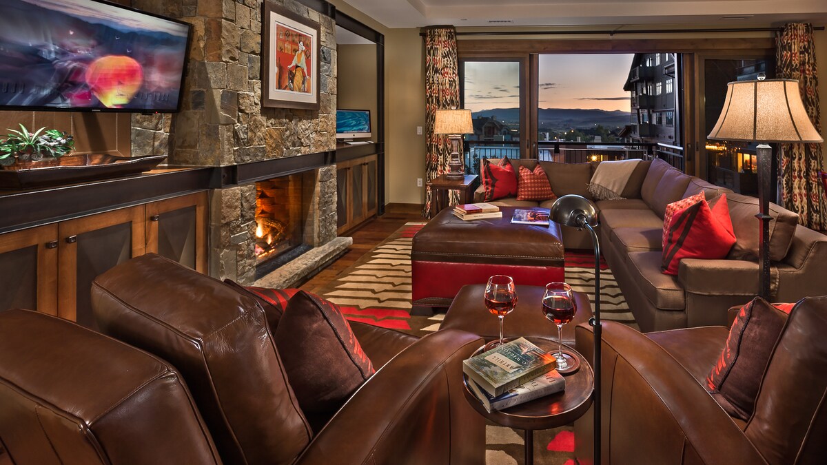 A spacious living area features a stone fireplace with a flat-screen TV above. Cozy seating includes a large sectional sofa adorned with decorative pillows, and two leather armchairs create an inviting space. Large windows provide views of the sunset and outdoor surroundings.